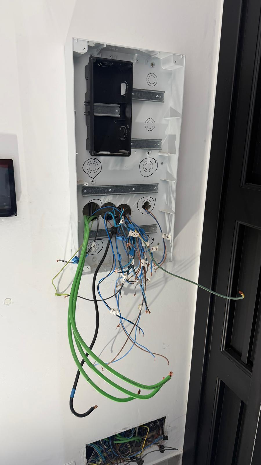 Close-up of the wall recess after strip-out — the original back-box and DIN-rail mounting plate are still in place because they are mechanically sound and reusable, while every wiring tail has been pulled clear, identified by circuit, and labelled with WAGO connector caps for protection. Reusing a sound back-box and mounting plate is the right call: it avoids opening up the wall surface around the recess, which would otherwise mean filling, sealing and repainting on a finished villa where the surrounding wall is in perfect condition. The 16 mm² green earth conductor and the supply tails are visible at the top, with the 18 outgoing circuit cables grouped below ready for connection to the new Chint board.