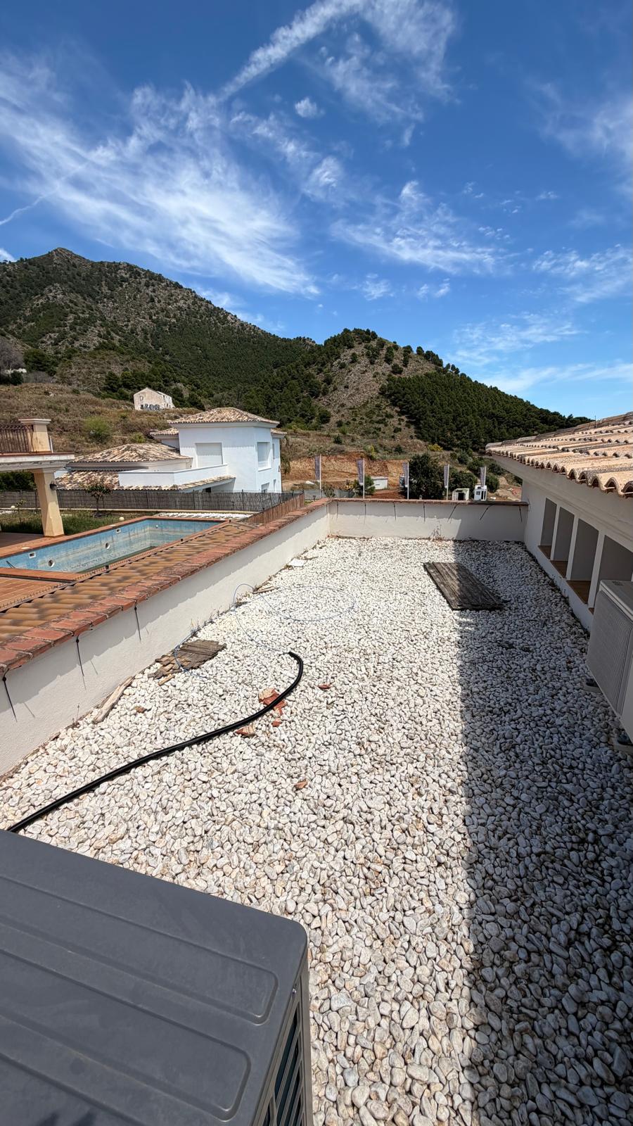 La zona de cubierta plana de grava junto a la villa principal — la unidad exterior del aire acondicionado (primer plano) es una de las cargas de alto consumo que justificó la elección de un inversor de 10 kW y una batería de 10 kWh. La piscina es visible a la izquierda con la Sierra de Mijas al fondo, ilustrando el microclima que hace de esta ubicación un lugar ideal para la generación solar durante todo el año.