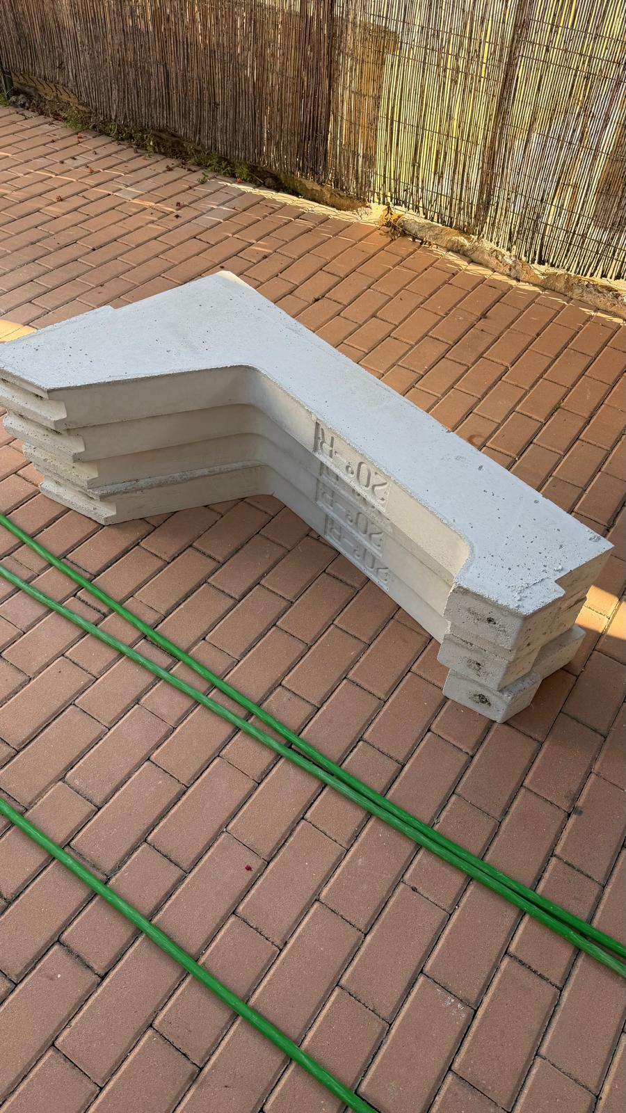 Escuadras de lastre de hormigón en forma de L utilizadas para montar paneles en la sección de cubierta plana de grava sin perforar — cuatro unidades apiladas, cada una marcada con '20°' indicando el ángulo de inclinación que proporcionan al panel. Con una inclinación de 20° en una cubierta plana a la latitud de Mijas, la penalización de rendimiento respecto a una superficie inclinada óptimamente hacia el sur es despreciable, mientras que el enfoque de lastre elimina cualquier riesgo de impermeabilización de cubierta.