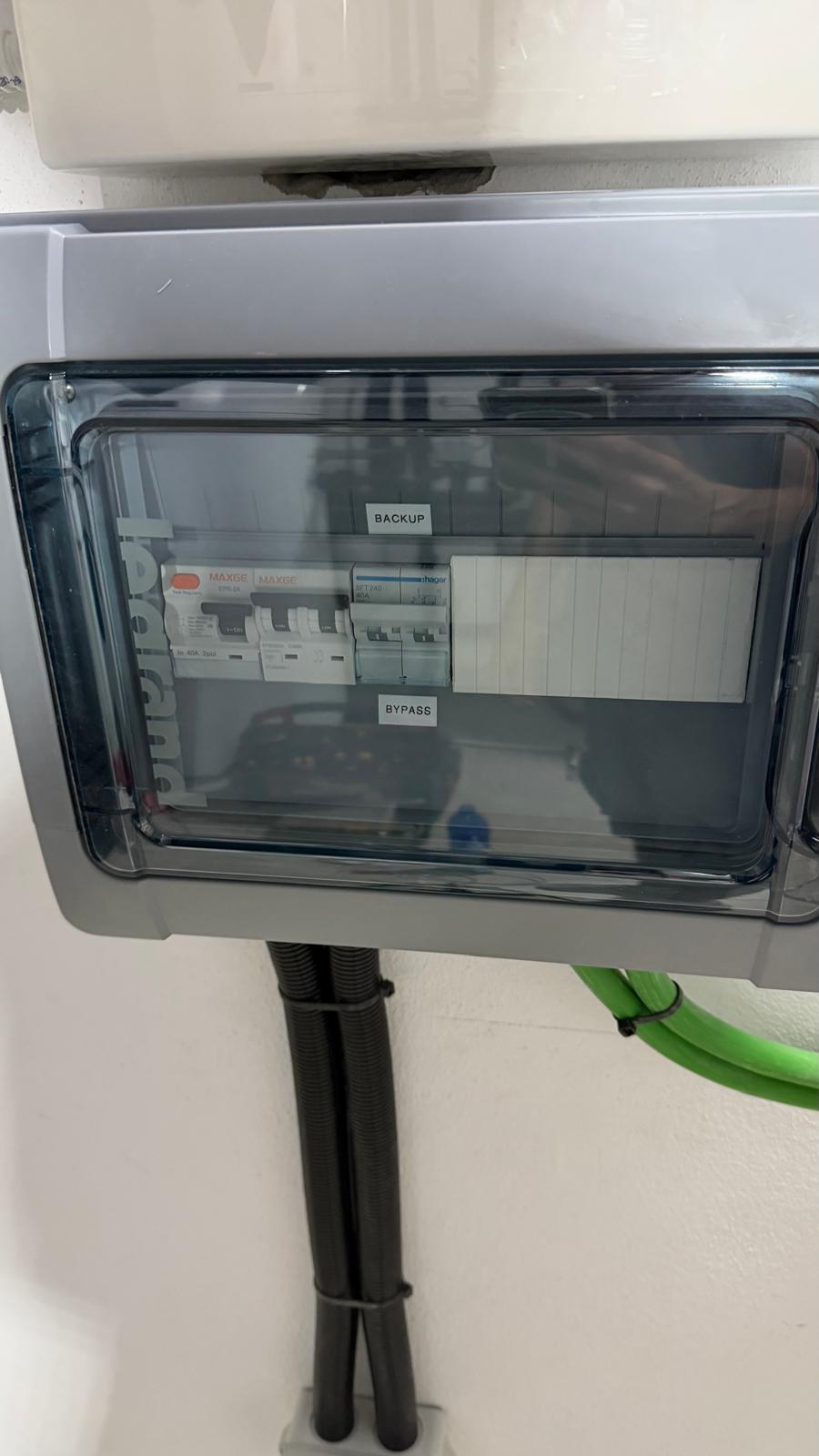 Subcuadro de carga crítica con tapa de acrílico transparente — la fila BACKUP (arriba) contiene los MCB para los circuitos respaldados por el inversor: alarma/videovigilancia, router, controlador de filtración de piscina, iluminación clave y nevera. La fila BYPASS (abajo) proporciona un bypass manual que permite conmutar esos circuitos directamente al suministro principal de red si el inversor requiere mantenimiento. Este etiquetado garantiza que cualquier electricista pueda entender la distribución del panel de un vistazo.