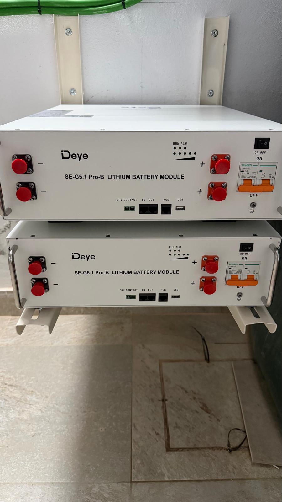 Los dos módulos de batería de litio Deye SE-G5.1 Pro-B en su rack apilado — 5,12 kWh cada uno, 10,24 kWh de capacidad utilizable total. Cada módulo tiene su propio interruptor DC de 100 A, puertos de comunicación por contacto seco y CAN bus, y un BMS que detecta automáticamente módulos adicionales si se añade una tercera unidad más adelante. Ambos están encendidos y listos para la puesta en marcha.