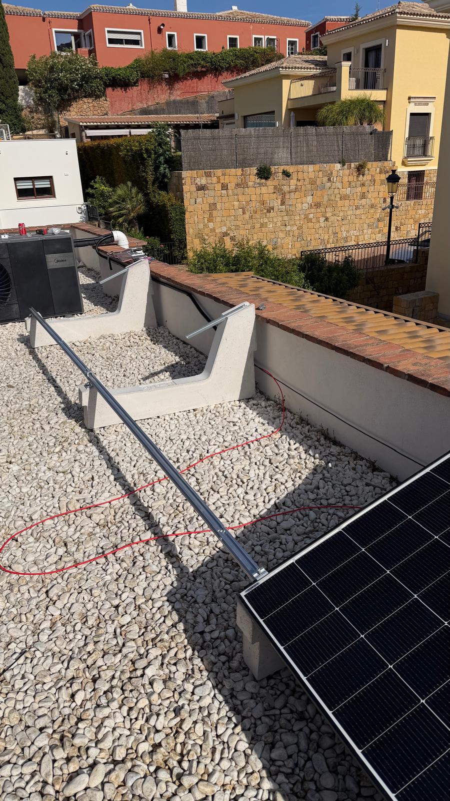 Un panel instalado en la sección de cubierta plana de grava usando bases de lastre de hormigón — no se requieren perforaciones de cubierta en esta sección, preservando la membrana impermeabilizante. El cable solar DC rojo está encaminado por la grava hasta el punto de penetración de cubierta. Las unidades exteriores del aire acondicionado son visibles a la izquierda — estas son algunas de las cargas de alto consumo que hicieron que el almacenamiento en batería fuera esencial para esta instalación.