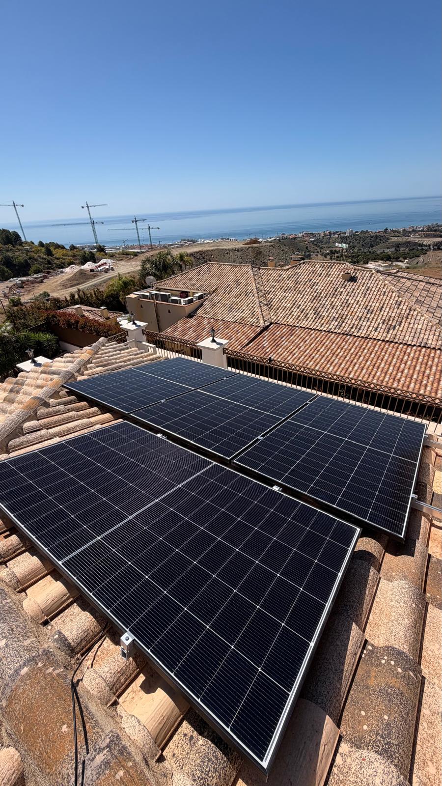 Cuatro paneles de 500 W completamente instalados y sujetos en el plano orientado al sur — grúas de construcción en la costa de Mijas visibles al fondo. Los paneles están cableados en dos strings de seis, cada uno alimentando una de las entradas MPPT duales del inversor Deye. El cable solar DC está encaminado por la hilera de tejas hasta el inversor abajo.
