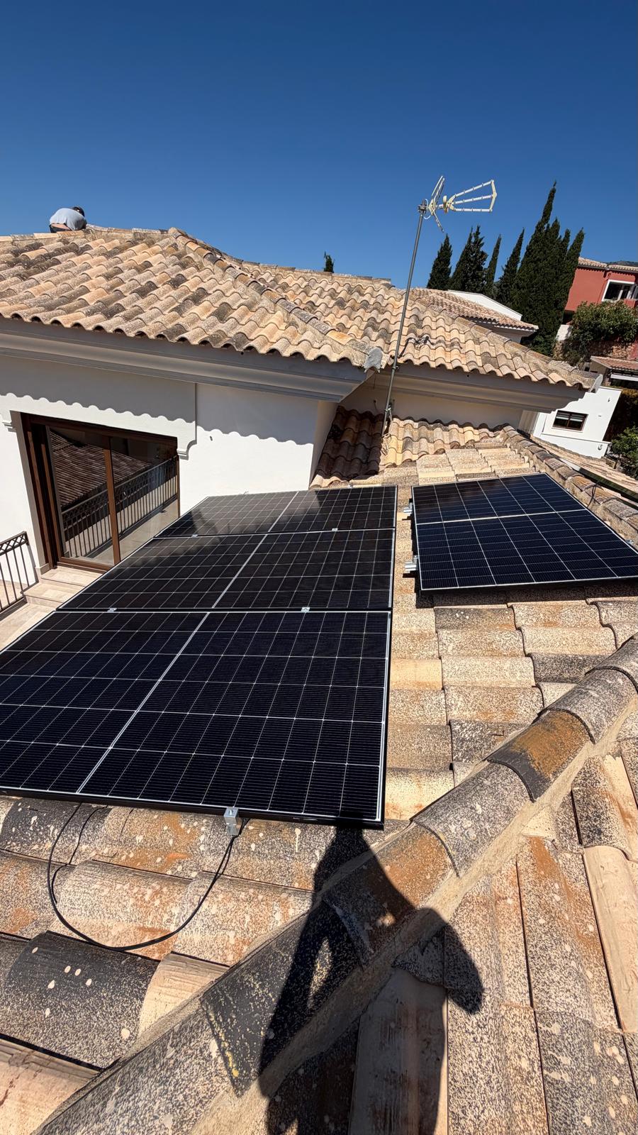 Paneles siendo posicionados en la cubierta orientada al sur — un técnico de LIT Electricians visible en la sección de cubierta superior mientras cuatro paneles ya están fijados en la sección inferior. Los tendidos de cable solar DC son visibles entre las dos secciones. El mástil de la antena de TV a la izquierda confirmó ausencia de sombras en ambos planos de cubierta durante el levantamiento previo a la instalación.