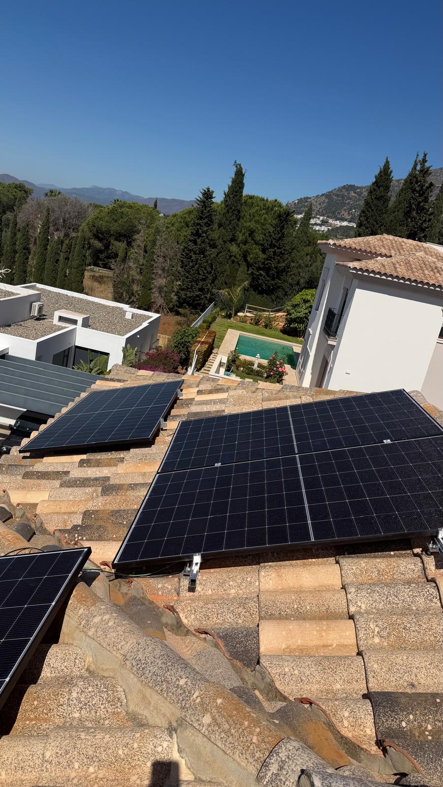 Vista desde la cumbrera mirando hacia el plano orientado al sur — cuatro paneles instalados en la sección principal con la piscina de la villa, jardín maduro y la Sierra de Mijas elevándose detrás. Esta perspectiva a nivel de cumbrera confirma la orientación sur limpia y la ausencia de auto-sombra entre las dos secciones de cubierta en cualquier momento del día.