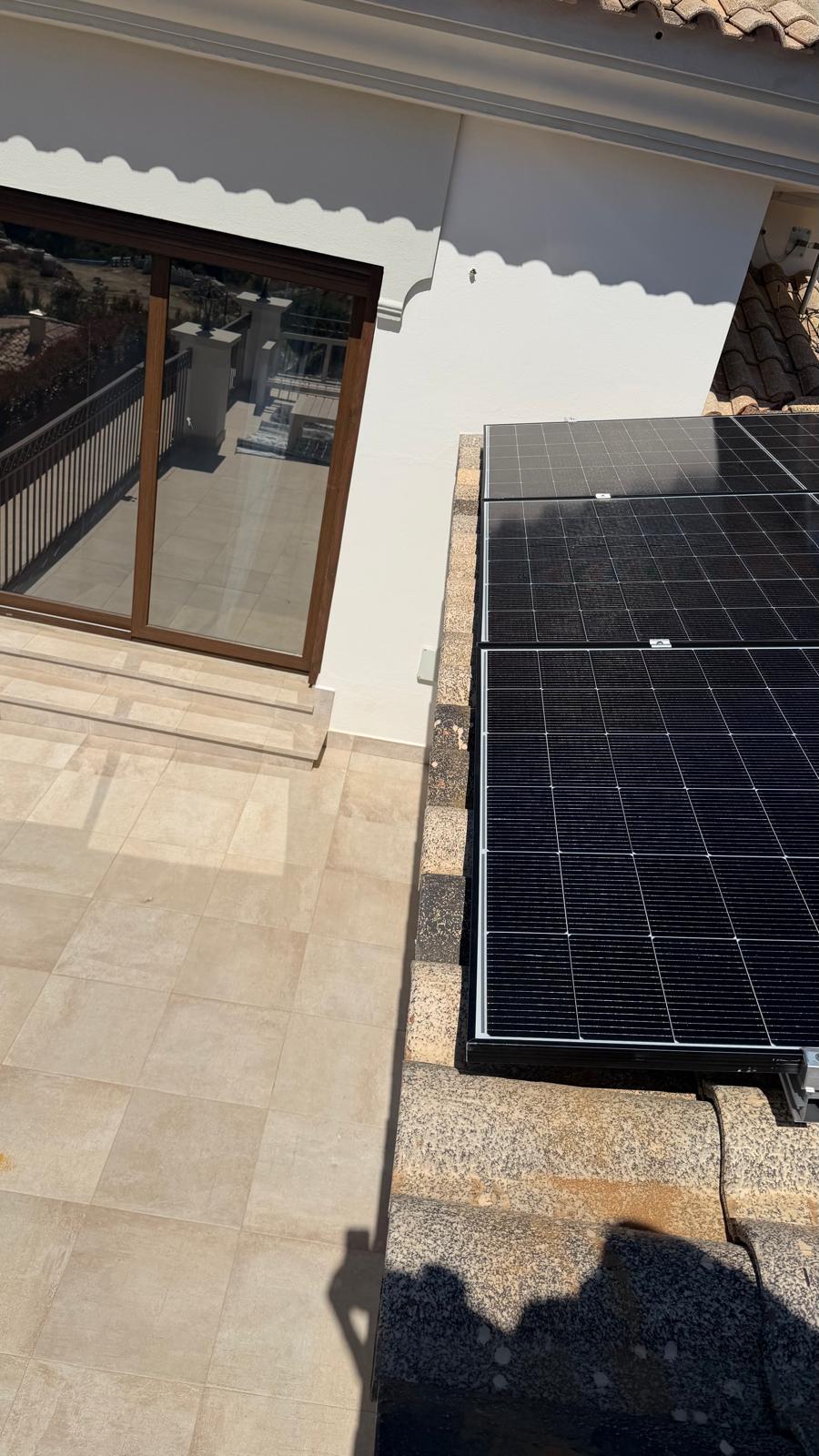 Vista desde la terraza superior mirando hacia abajo a dos paneles completados en la sección de cubierta inferior — los paneles encajan a ras en el sistema de raíles de aluminio, con abrazaderas centrales y de extremo visibles en los bordes del marco del panel. El cable DC sale por la esquina inferior y se canaliza en tubo solar hasta el inversor.