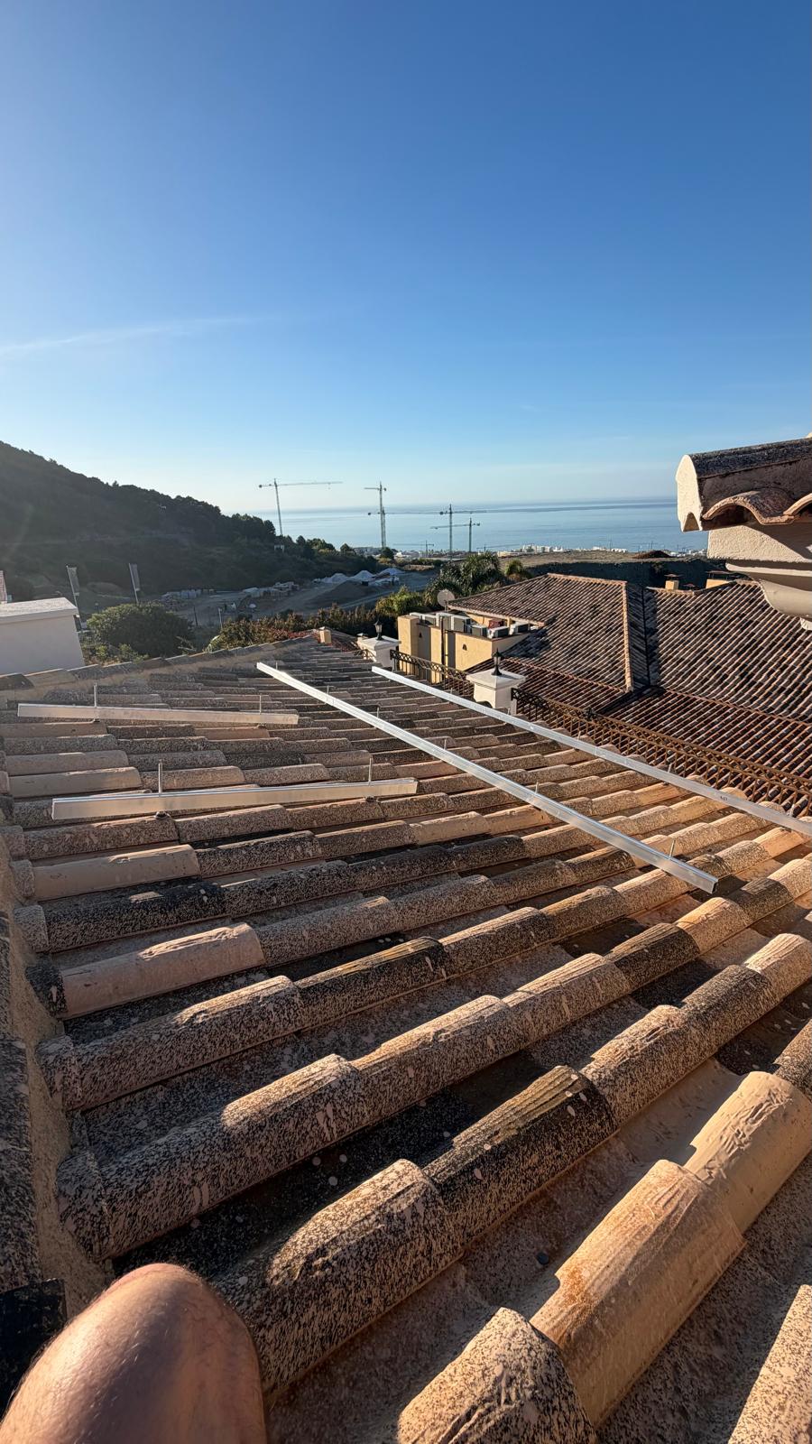 Vista a la hora dorada desde la cubierta al inicio de la instalación — el primer raíl de montaje tendido a lo largo de la hilera de tejas, con el mar Mediterráneo y la costa del Sol visible con la luz de la mañana temprana. Esta orientación y elevación (colinas de Mijas, ~36,6° N de latitud) entrega un estimado de 1.500 kWh por kWp al año.
