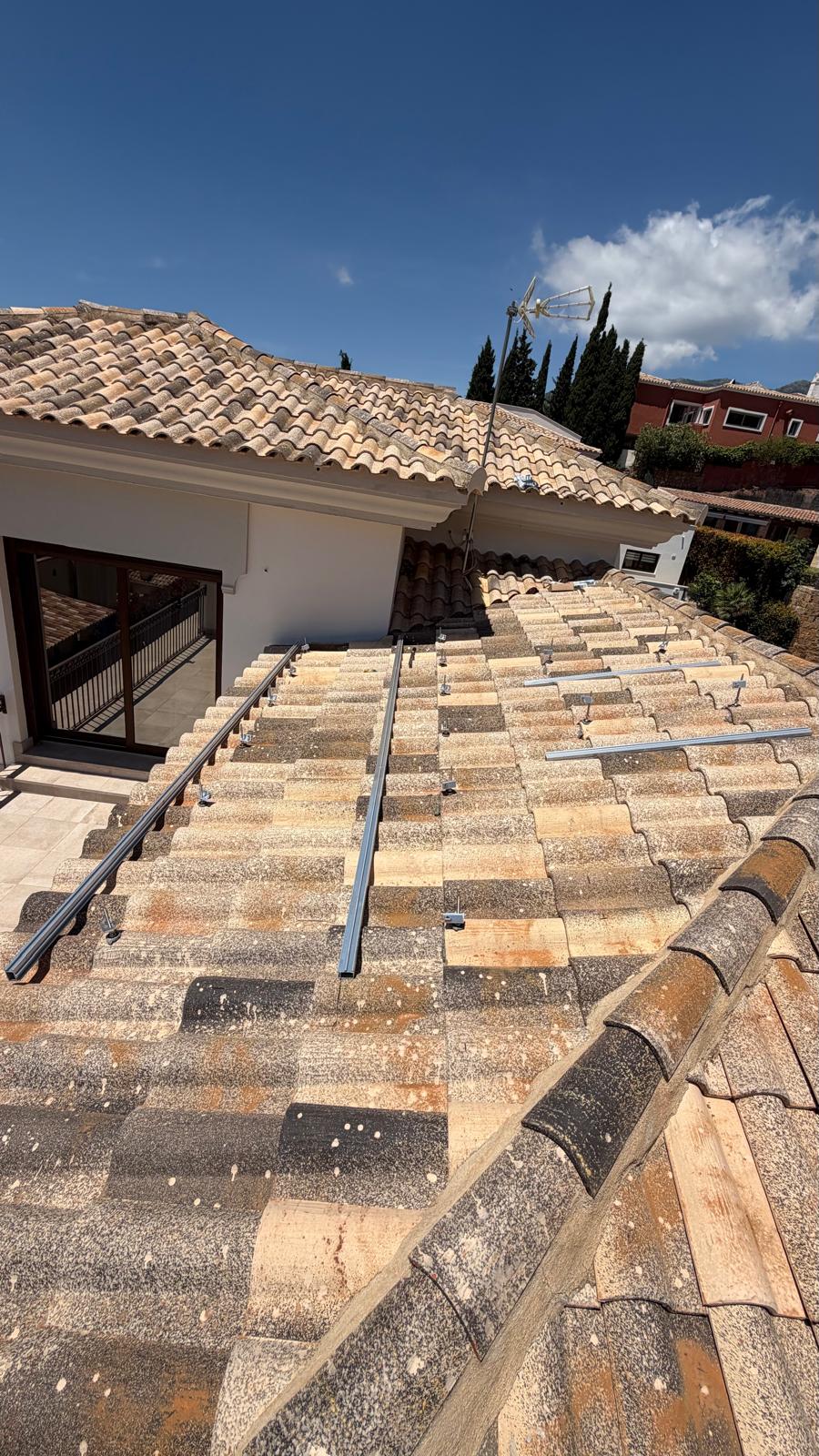 Primer plano de la instalación de raíles de montaje en la cubierta de tejas de terracota orientada al sur — dos raíles de aluminio fijados con ganchos pasarela, alineados y nivelados antes de colocar los paneles. La terraza y las puertas correderas de la villa son visibles al fondo.