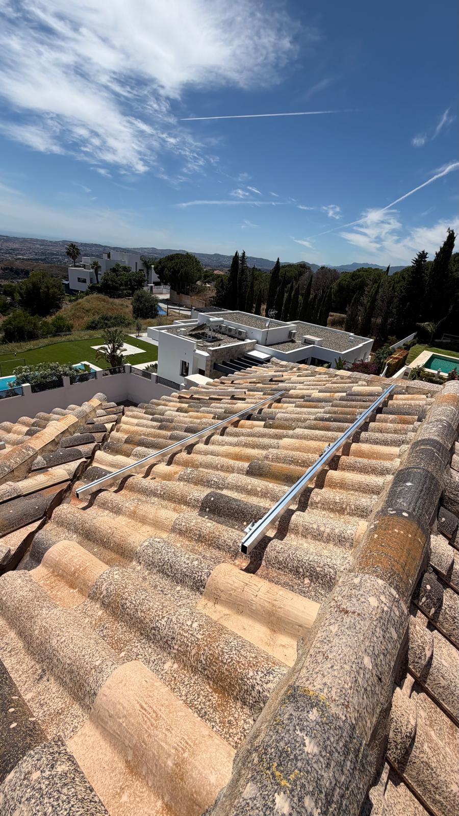 Raíles de montaje instalados en la sección secundaria de cubierta, con un amplio panorama interior de las colinas de Mijas y villas vecinas abajo — la piscina de la propiedad adyacente visible abajo a la izquierda. Este plano de cubierta utiliza una entrada MPPT separada en el inversor Deye, pre-puesta en marcha y lista para recibir el string de ampliación de paneles futura sin ningún cambio en el inversor ni la documentación de red.