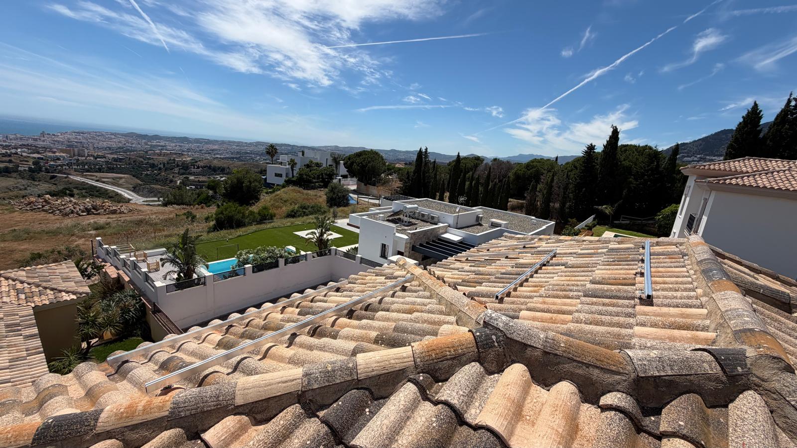 Vista panorámica desde la cubierta de la villa de Mijas durante la instalación de los raíles de montaje — tres raíles de aluminio tendidos sobre las tejas de terracota españolas, con la piscina y villas vecinas visibles abajo y la costa de Málaga extendiéndose hasta el horizonte.