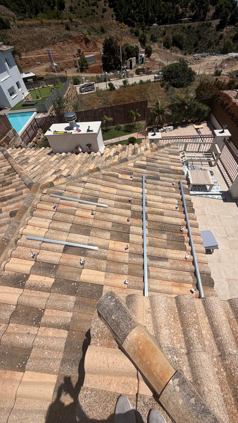 Vista aérea desde la cumbrera mirando hacia el plano sur de la cubierta — raíles de montaje instalados en toda el área de paneles con ganchos pasarela visibles a intervalos regulares. La piscina, el jardín y la ladera de Mijas son claramente visibles abajo, confirmando el emplazamiento elevado orientado al sur sin obstáculos de sombra en el plano principal.