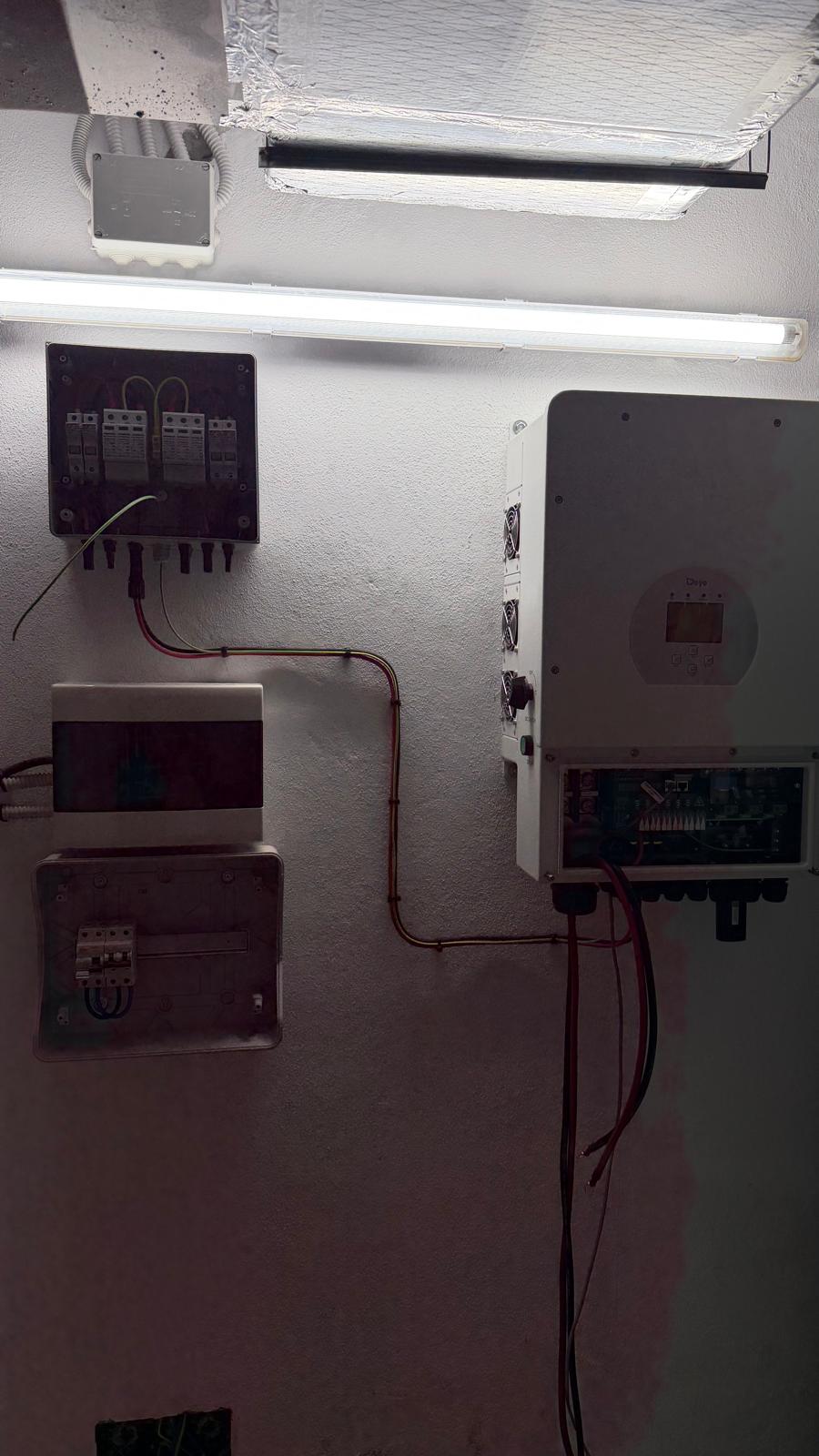 Vista general del cuarto técnico con el conjunto completo de protecciones CC y CA: caja combinadora con protección contra sobretensiones DC (arriba a la izquierda) con descargadores tipo 1+2, dos cuadros de distribución (centro izquierda) — el superior para el suministro principal, el inferior para el subcuadro de carga crítica — y el inversor Deye (derecha) con el cableado de batería y red en rojo y negro.