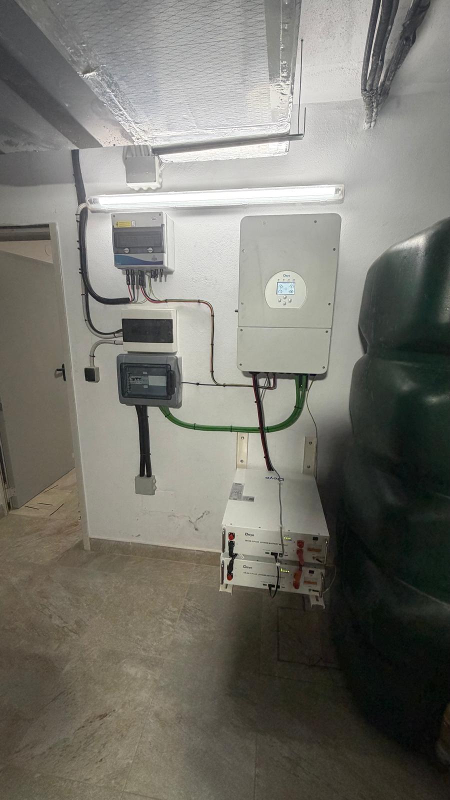 Vista general completa del cuarto técnico de la instalación terminada — caja combinadora de protección DC (arriba izquierda) con descargadores tipo 1+2, contador de red, subcuadro de carga crítica (columna izquierda), inversor Deye SUN-10K-SG04LP1 (centro), seccionador DC (centro-derecha) y 2 módulos de batería Deye SE-G5.1 Pro-B en su rack de suelo (abajo derecha). El cableado de tierra verde y el cableado DC de batería rojo/negro están completamente ordenados y etiquetados.