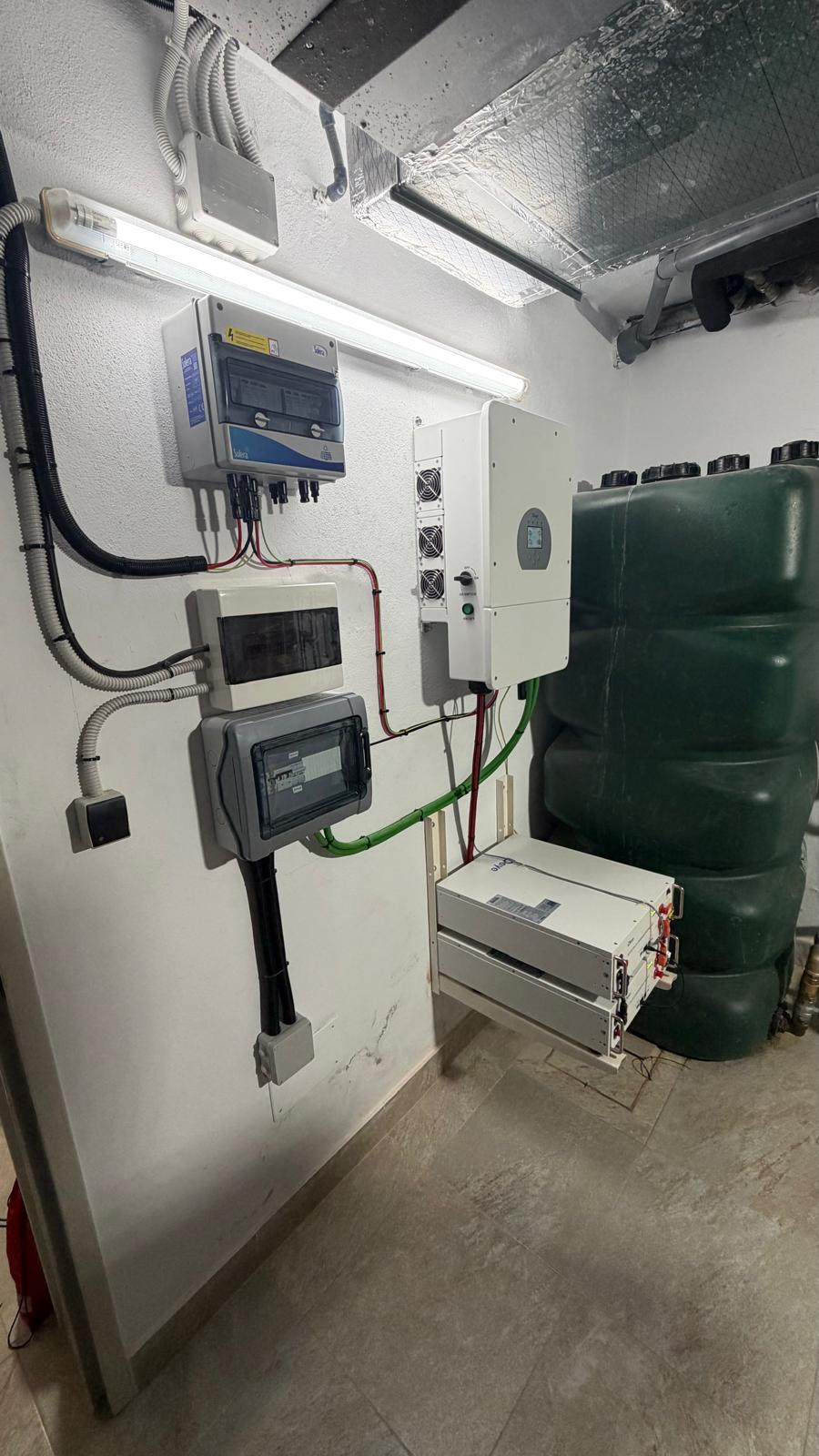 Vista gran angular del cuarto técnico terminado — el sistema completo de izquierda a derecha: combinador DC con protección contra sobretensiones, inversor Deye con ventiladores de refrigeración visibles, cuadros de distribución y el rack de baterías a nivel de suelo. El depósito de almacenamiento de agua verde a la derecha confirma que este es el cuarto de instalaciones principal de la villa. El sistema fue puesto en marcha y verificado el 22 de mayo de 2026.