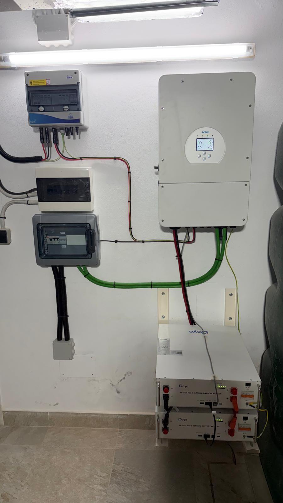 Cuarto técnico terminado — inversor Deye SUN-10K-SG04LP1 (arriba derecha) con combinador de protección DC (arriba izquierda), contador de red y subcuadro de carga crítica (columna izquierda), y 2 módulos de batería de litio Deye SE-G5.1 Pro-B en su rack mural (abajo derecha). Los cables de tierra verde y los cables de batería DC rojo/negro están canalizados ordenadamente.