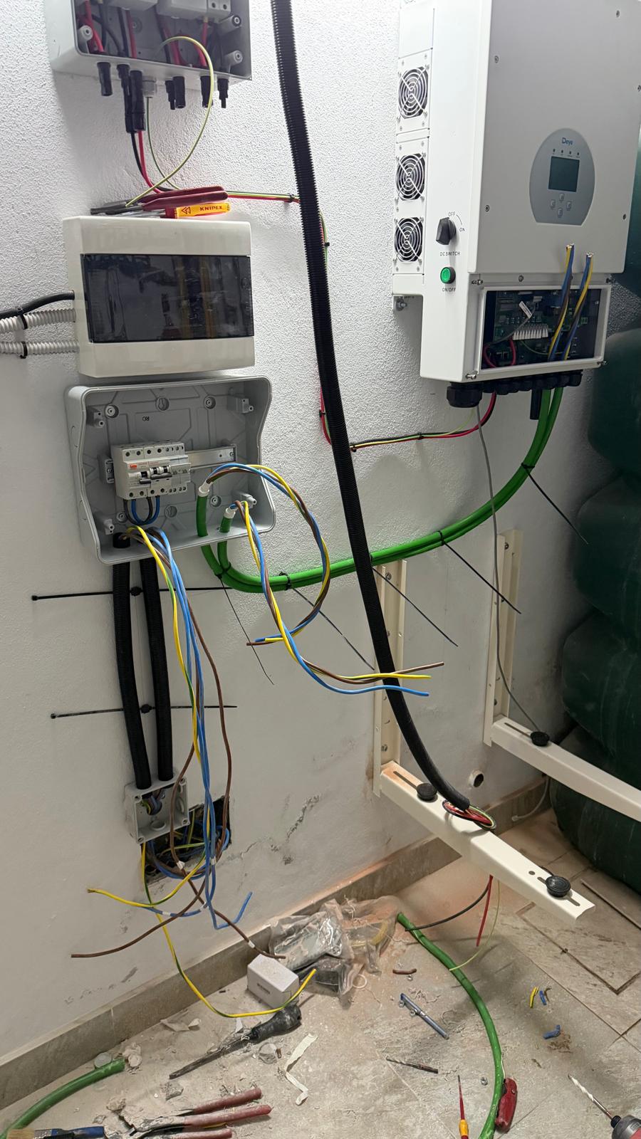Cuarto técnico a mitad de la instalación — cables solares DC desde la cubierta entrando en la caja combinadora (arriba izquierda), cuadro de distribución principal y subcuadro de carga crítica siendo cableados (centro izquierda) y el inversor Deye con su bahía de conexiones abierta (derecha). Los cables de tierra verde, las acometidas DC solares negras y los cables CA multiconductor son visibles siendo ordenados antes de la terminación final.