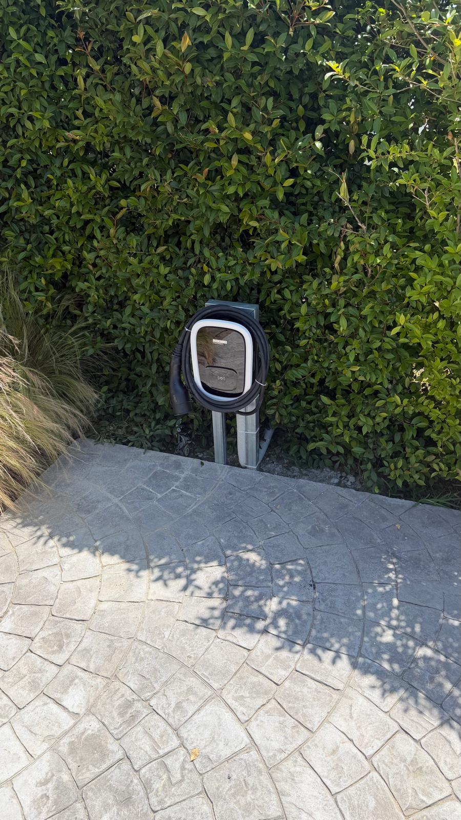 Wide view of the completed install in the garden of the San Pedro de Alcántara villa — the Orbis Viaris Uni 7.4 kW pedestal sits in front of the laurel hedge at the head of the stamped-concrete parking bay, where it can serve either parking position with the 5 m Type 2 tether without any cable crossing the open driveway. The galvanised pedestal anchor plate is bolted through the stamped-concrete slab into chemical anchors set into the underlying concrete; the buried supply trench runs from this point back across the driveway to the technical room on the side wall of the villa where the consumer unit and the new dedicated 2-pole 40 A Type A + 6 mA DC RCBO are housed. The bright zinc finish of the pedestal will weather to a uniform matte grey within twelve months and read as garden ironwork rather than as electrical equipment — the design intent on a residential property in San Pedro de Alcántara where visual integration with the garden mattered to the client.