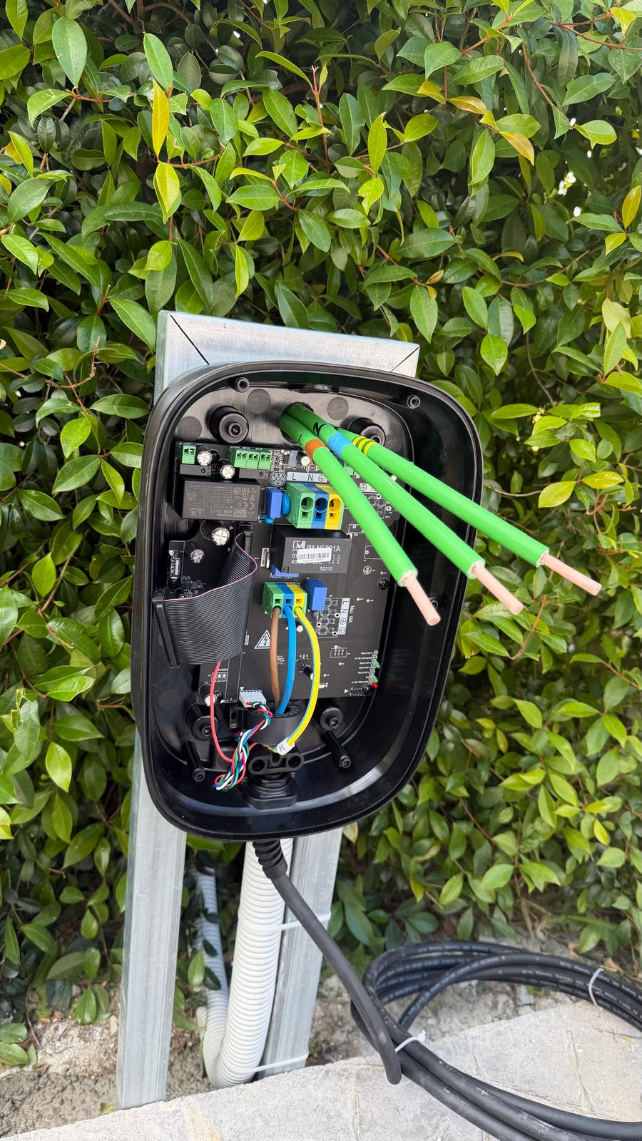 Orbis Viaris Uni 7.4 kW with the front cover removed during commissioning at this San Pedro de Alcántara villa — the 3G6 mm² RZ1-K (AS) supply core lands on the L (line) terminal, with neutral on the blue terminal and the protective earth on the green-and-yellow terminal, all entering through the IP67 polyamide gland visible at the bottom of the enclosure. The internal contactor module is the dual-rated 32 A device that switches the line and neutral together; the small DC residual current sensor inside the contactor block is the 6 mA detector that ITC-BT-52 makes mandatory on EV charging circuits. The yellow conductor and ribbon harness running to the lower-left PCB carry the CP/PP control-pilot and proximity-pilot signals back to the on-board EV charger via the Type 2 plug. The white corrugated conduit visible at the bottom runs inside the hollow galvanised pedestal back to the consumer unit on the villa wall.