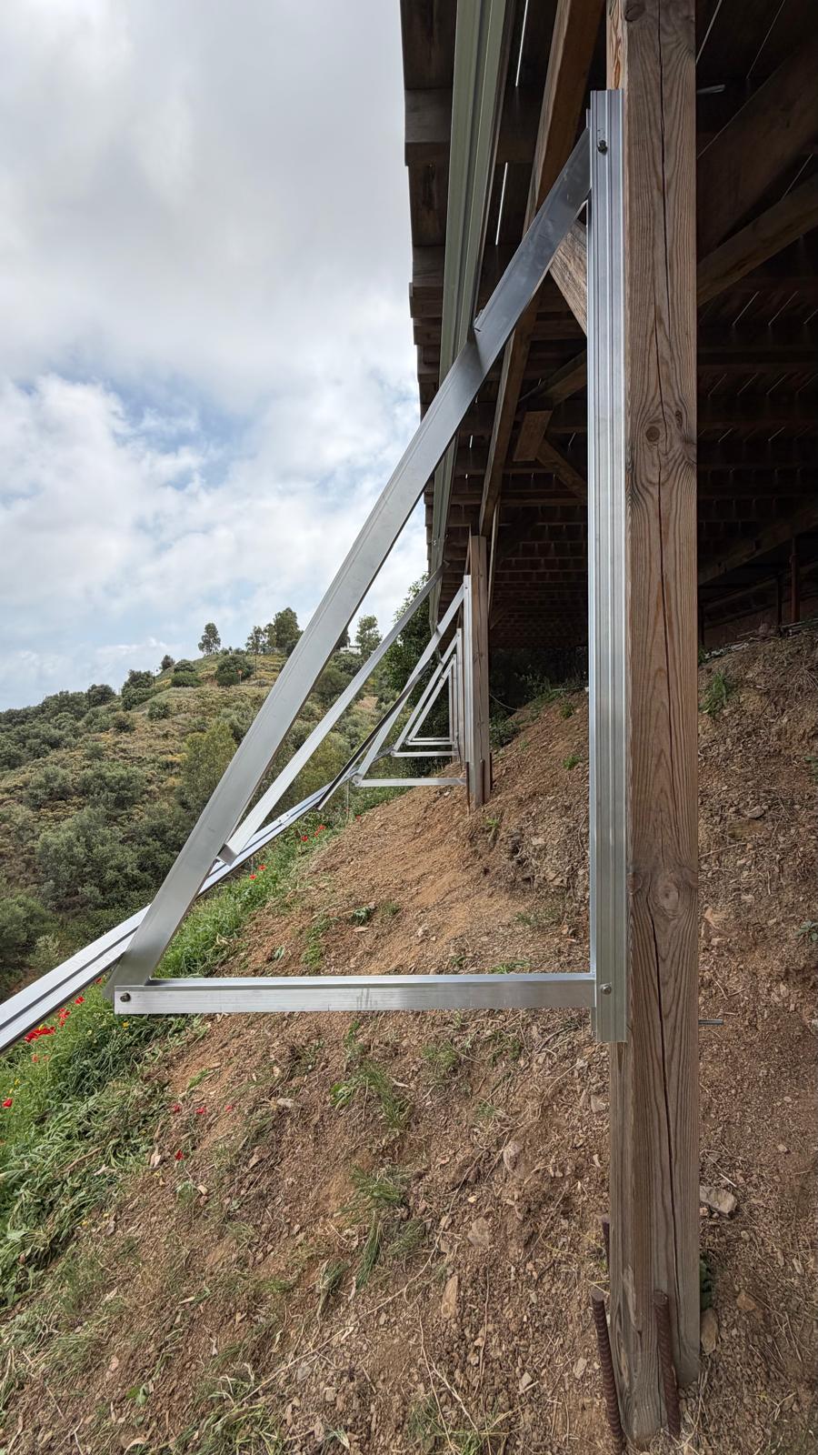 Detalle del triángulo A de acero galvanizado — obsérvese el tirante diagonal que va desde el punto alto del plano de panel hasta un anclaje a suelo independiente por delante de las columnas de la terraza, derivando la carga de succión por viento desde las fijaciones superiores atornilladas a la columna hacia la roca madre. El acabado brillante es el recubrimiento de zinc del baño de galvanización en caliente, que sella cada corte, cada perforación y cada soldadura por dentro y por fuera — algo que la pintura o el termoesmaltado en polvo no consiguen en los extremos cortados.