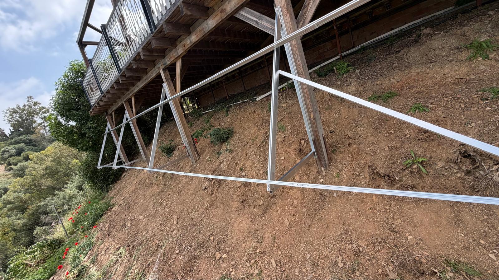 Fase de construcción de la estructura — triángulos A a medida de acero al carbono galvanizado en caliente de 60×60 mm atornillados a las columnas existentes de hormigón armado de la terraza por arriba y a anclajes químicos introducidos 250 mm en la roca madre por abajo. Tres triángulos A cubren los 6,5 m de longitud previstos para la instalación, con los tirantes diagonales sacando el plano de panel por delante del vuelo de la terraza, hacia el cielo despejado. La galvanización en caliente conforme a EN ISO 1461 aporta un recubrimiento de zinc de 70–85 µm con vida útil de diseño >50 años en este emplazamiento interior de Mijas con corrosividad C2.