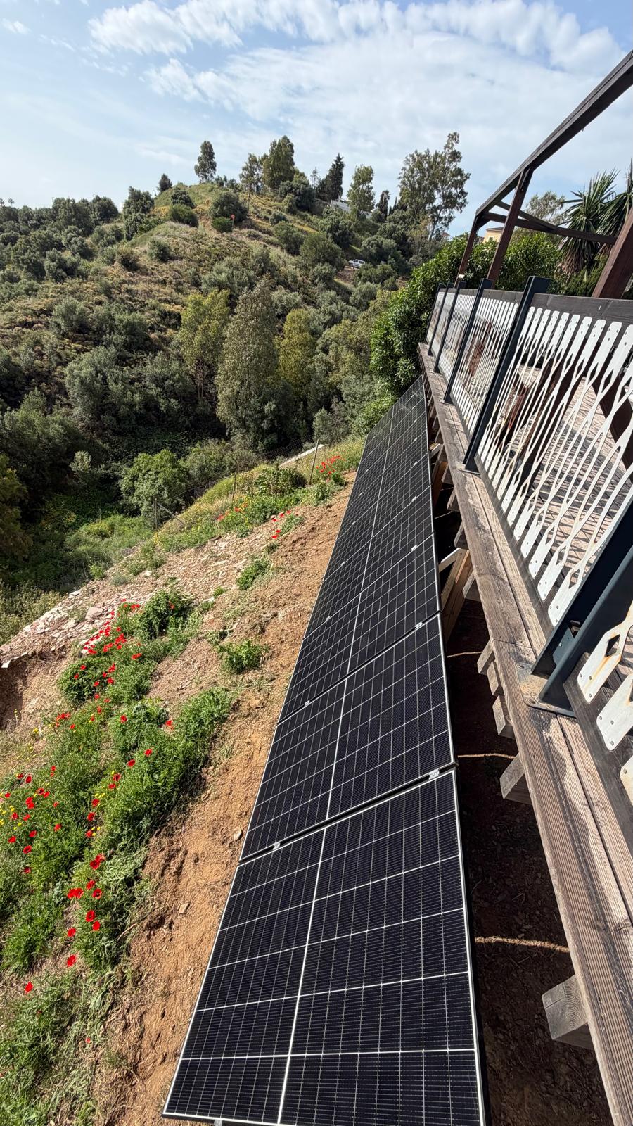 Vista en picado de la hilera de paneles desde la terraza superior — línea larga y uniforme de seis paneles mono PERC idénticos de 500 W sujetos al carril de aluminio superior, todos con inclinación consistente y con el horizonte suroeste libre de obstrucciones por delante. El acero brillante de los tirantes diagonales inferiores se ve pasando por debajo de los paneles y anclándose en la ladera. Esta perspectiva cenital muestra la geometría de producción que aporta la orientación suroeste: sol al final de la mañana, toda la tarde y al inicio de la noche sin que los paneles se sombreen entre sí.