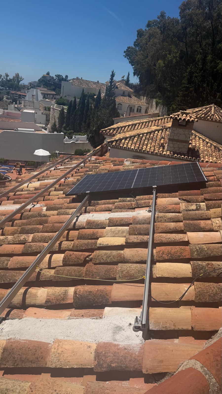 Sistema de raíles de aluminio instalado sobre las tejas árabes envejecidas — mordazas centrales y de extremo visibles. Arquitectura tradicional del pueblo blanco de Mijas Pueblo al fondo.